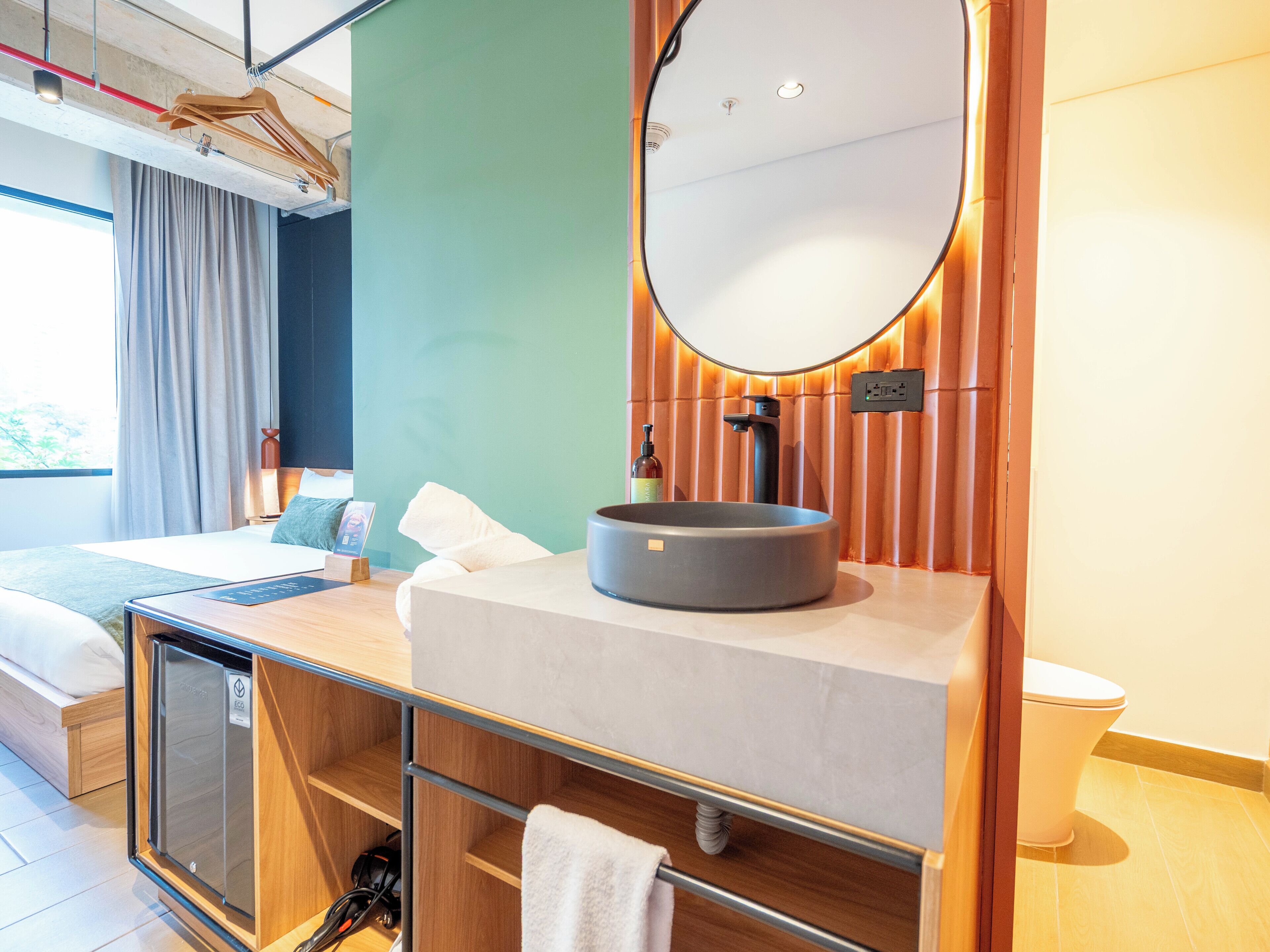Una habitación de hotel moderna con una cama, un espejo redondo, un lavabo y un mueble pequeño.