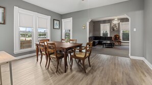 A dining area with a wooden table and chairs, a ceiling fan, and large windows with white blinds.