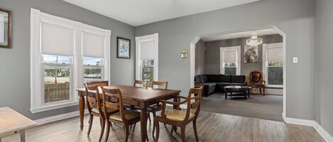 A dining area with a wooden table and chairs, a ceiling fan, and large windows with white blinds.