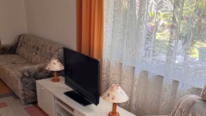 Een woonkamer met een zithoek, een televisie op een witte kast, twee lampen en een gordijn met een patroon.
