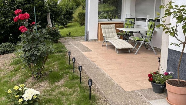 Een overdekt terras met een tafel en stoelen, omgeven door potplanten en een tuinpad.