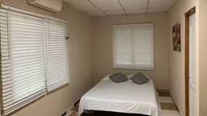 Een smalle hotelkamer met een enkel bed, witte muren en een raam met jaloezieën.