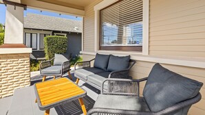 Une terrasse couverte, aménagée avec des meubles en osier, une table en bois et offrant une vue sur la maison voisine.