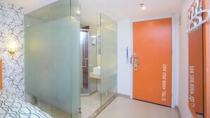 Een moderne hotelkamer met een glazen scheidingswand, een badkamer met douche, en een oranje deur met een telefoonnummer.