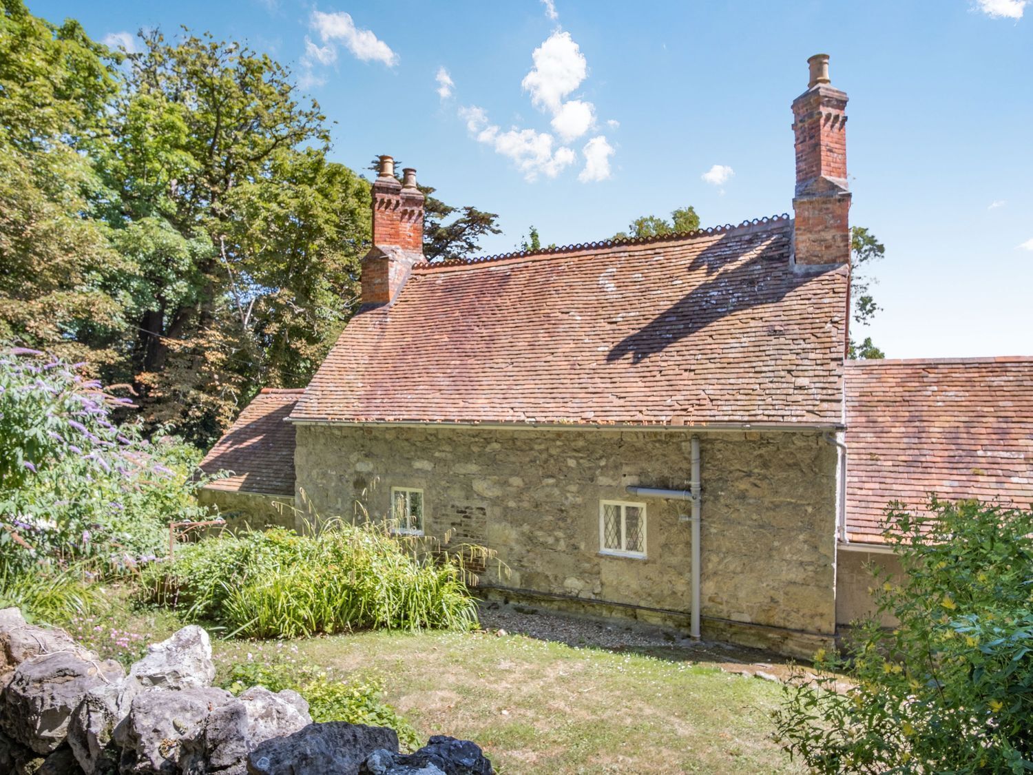A stone house with a tiled roof, surrounded by greenery and a stone wall.