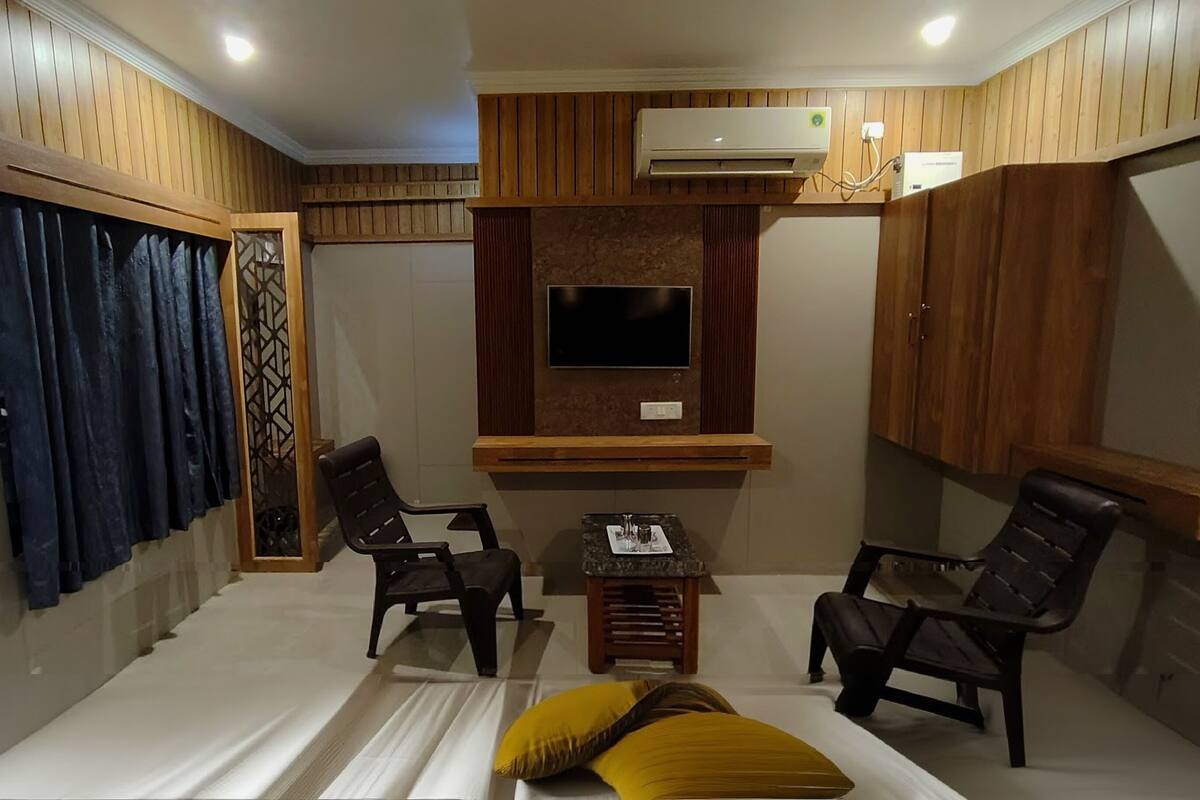 A room with a bed, two chairs, a small table, an air conditioning unit, and a TV mounted on the wall.