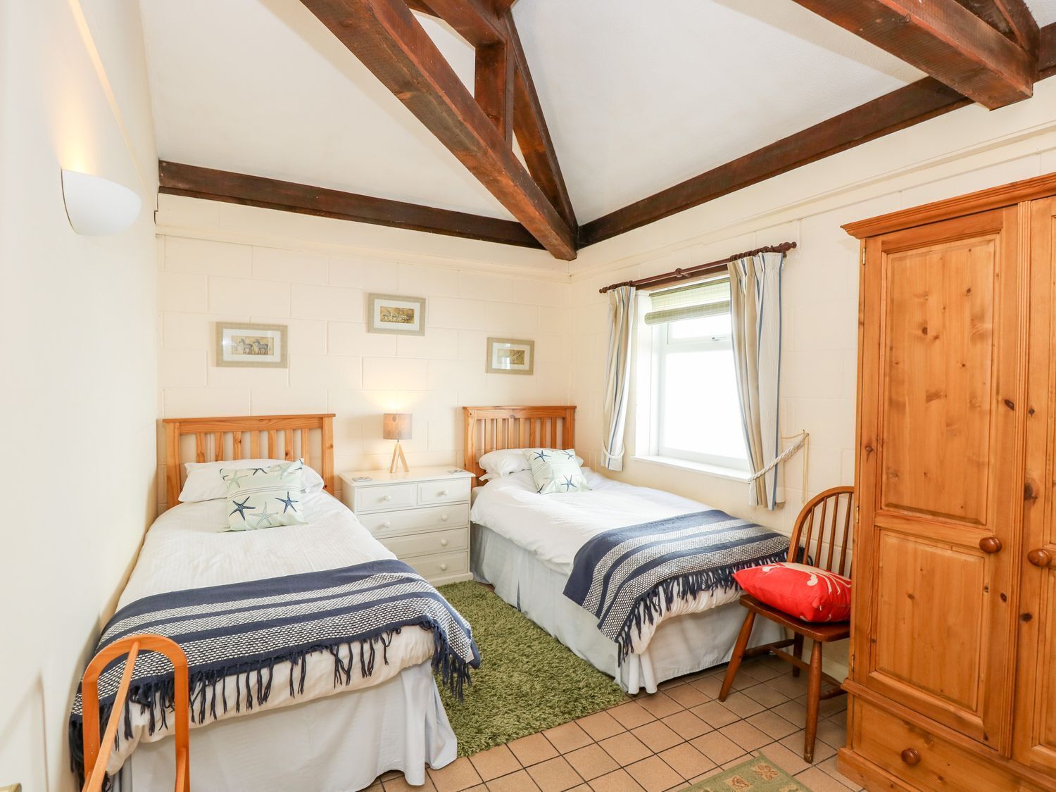 A bedroom with two beds, a wooden wardrobe, a window with curtains, and a green rug.