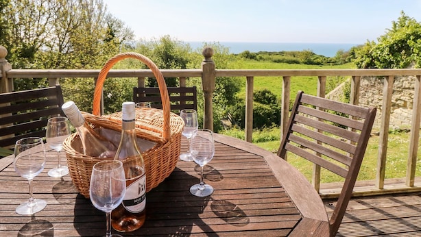 A wooden table with a wicker basket, wine bottles, and glasses on a balcony overlooking a green landscape and the sea. - Swallows (Ventnor)