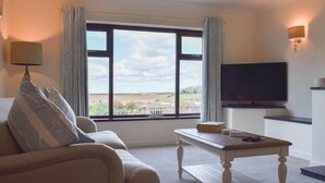 A living room with a sofa, a coffee table, a flat-screen TV, and a large window with a view of the countryside. - Cae Coch (Brynteg)