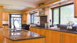 A modern kitchen with granite countertops, wooden cabinets, a stainless steel sink, and a large window with blinds. - Cae Coch (Brynteg)