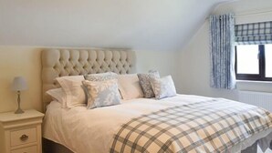 A bedroom with a tufted headboard, a bed with a plaid bedspread, a bedside table with a lamp, and a window with curtains. - Cae Coch (Brynteg)