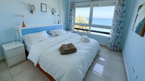 A bedroom with a large bed, a nightstand, a window with a view of the sea, and decorative wall art.