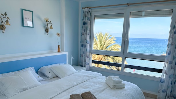 A bedroom with a bed, pillows, a bedside table, a wall-mounted light, a framed picture, and a window with a view of the sea.