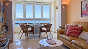A living room with a view of the ocean, a brown leather sofa, wicker chairs, a glass coffee table, and a small round table with a bowl and glasses.
