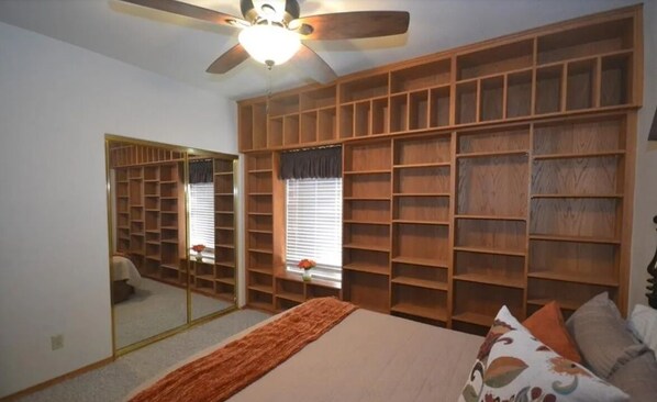 A bedroom with a large bed, a ceiling fan, a wardrobe with multiple shelves, and a mirror.