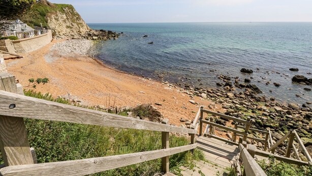A coastal view with a sandy beach, rocky shoreline, and a wooden staircase leading down to the beach. - The Lobster Pot (Ventnor)