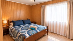 Un dormitorio con cabecera de madera, cama con colcha estampada, mesita de noche con lámpara y ventana con cortinas traslúcidas.