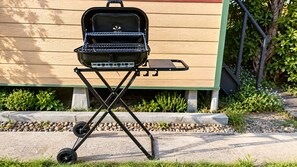 Un barbecue portable doté d’un support pliable, placé sur une surface en béton, avec une clôture en bois et de la verdure en arrière-plan.