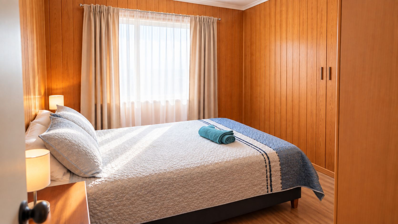 A neatly made bed with a quilt, pillows, and a rolled towel in a room with wooden walls and a window with curtains.