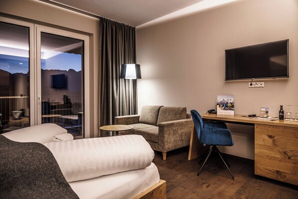 Quarto de hotel com cama, escrivaninha, cadeira, abajur e televisão.