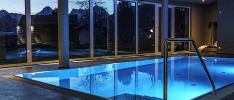 Una piscina interior moderna con paredes de vidrio transparente, una escalera metálica y vistas a montañas.