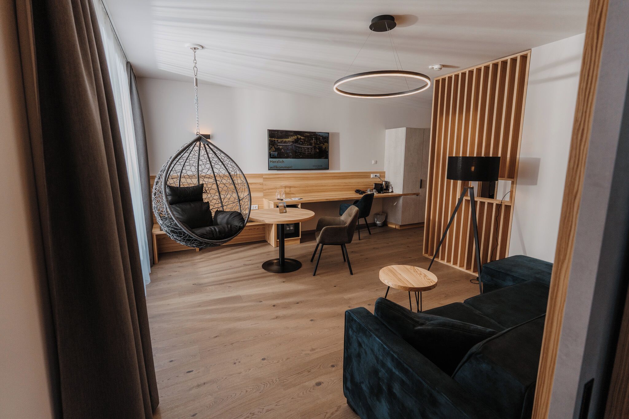 A modern interior with a hanging chair, a round wooden table, a dark sofa, and a wooden partition.