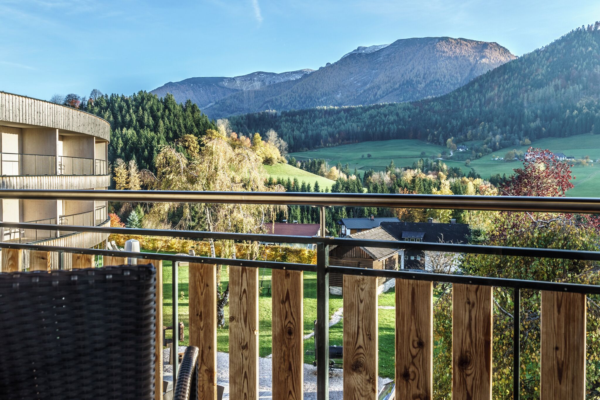 A balcony with a view of a mountainous landscape, a building with a wooden facade, and a grassy area with trees.