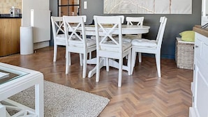 A dining area with a round table and white chairs, a clock on the wall, and a large abstract painting. - Cosy Sunny Apartment Near Zeedijk (Knokke-Heist)