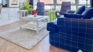 A modern living room with a blue sofa, white cabinets, a coffee table, and a view of buildings through large windows. - Cosy Sunny Apartment Near Zeedijk (Knokke-Heist)
