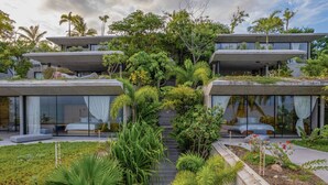 Un edificio moderno con varios niveles, amplios ventanales y un jardín exuberante con diversas plantas y un sendero que conduce a la entrada.