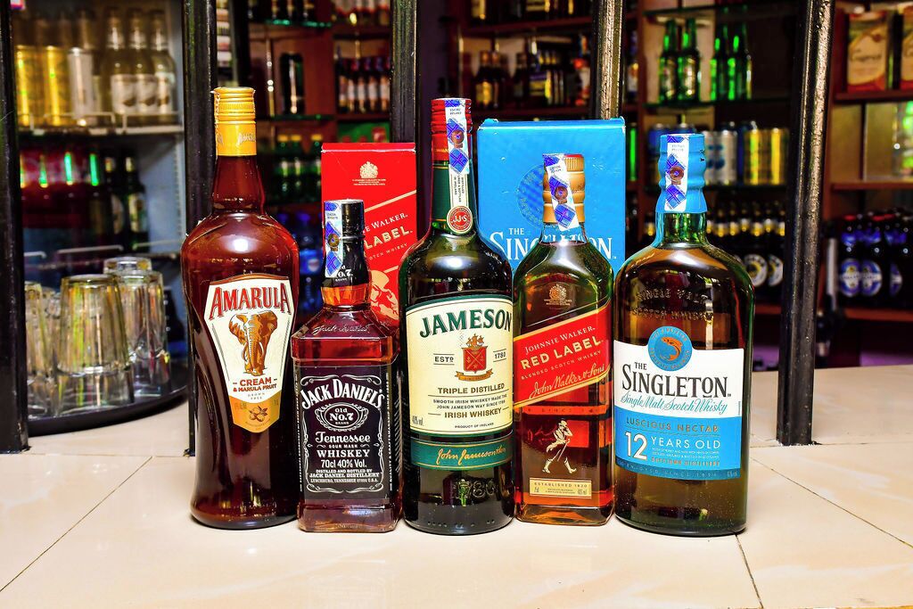 A selection of whiskey bottles on a bar counter with a background of shelves filled with various bottles.