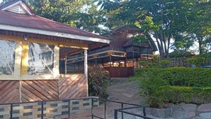 A wooden building with a corrugated metal roof, large windows, and a stone pathway leading to a wooden deck. - Rosa Gardens Hotel (Muhoroni)