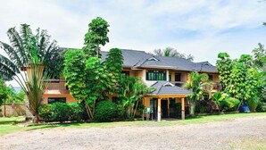 A two-story house with a covered porch, surrounded by lush greenery and palm trees. - Rosa Gardens Hotel (Muhoroni)