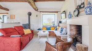 Un salon chaleureux avec un canapé rouge, un fauteuil en cuir marron et une table basse blanche. On y trouve une cheminée ornée de poteries bleues et blanches, ainsi qu’une fenêtre donnant sur l’extérieur.