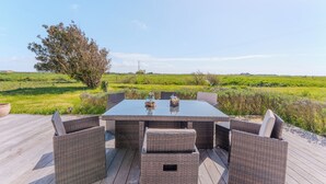 Un espace repas extérieur avec une table à plateau de verre et des chaises en osier, entouré d’une terrasse en bois et offrant une vue sur un champ verdoyant.