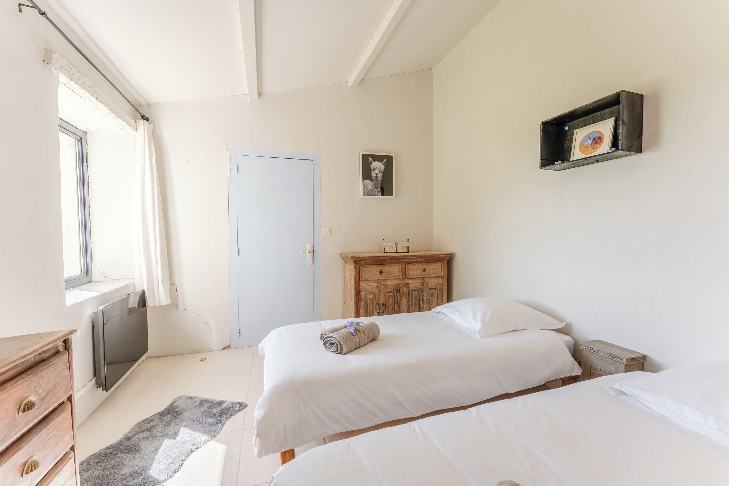 A bedroom with two beds, a wooden dresser, a small cabinet, a framed picture on the wall, and a window with curtains.
