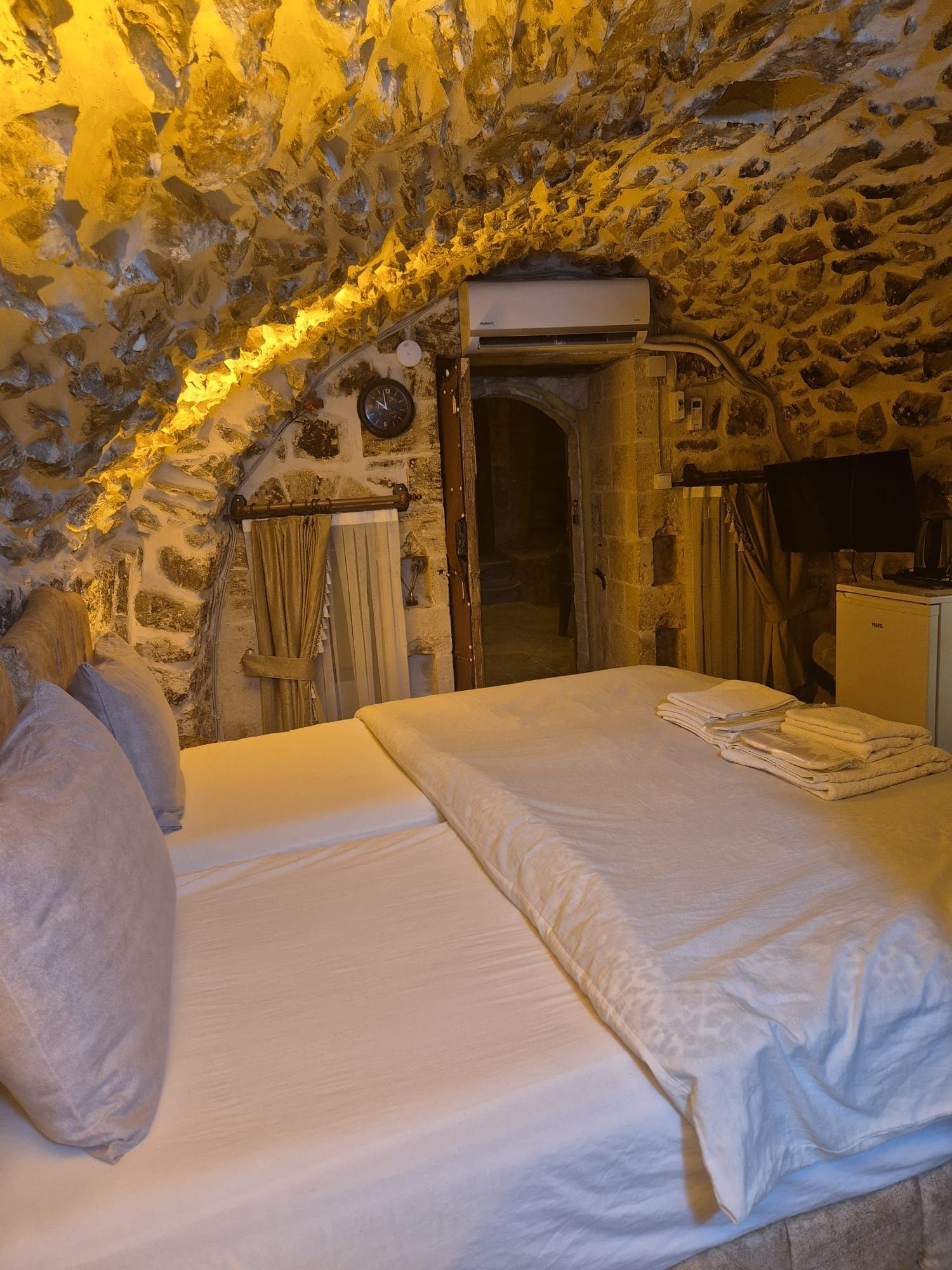 Una habitación con techo de piedra, una cama con ropa blanca, un reloj y un escritorio pequeño con un ordenador.
