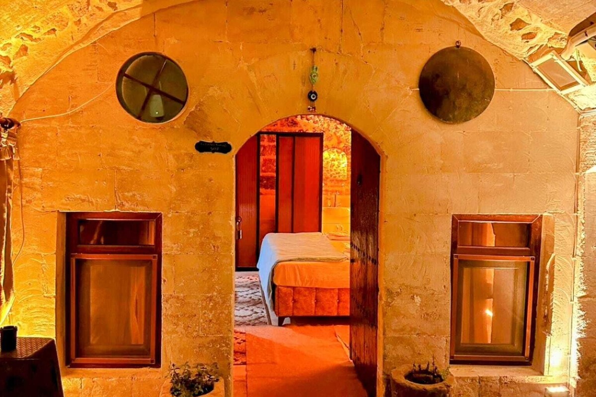A room with a stone archway leading to a bed with a wooden headboard, a small circular window, and a door.
