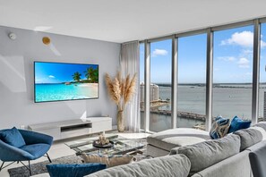 Una sala de estar moderna con un televisor grande, asientos cómodos, una mesa de centro y vistas al agua y edificios.