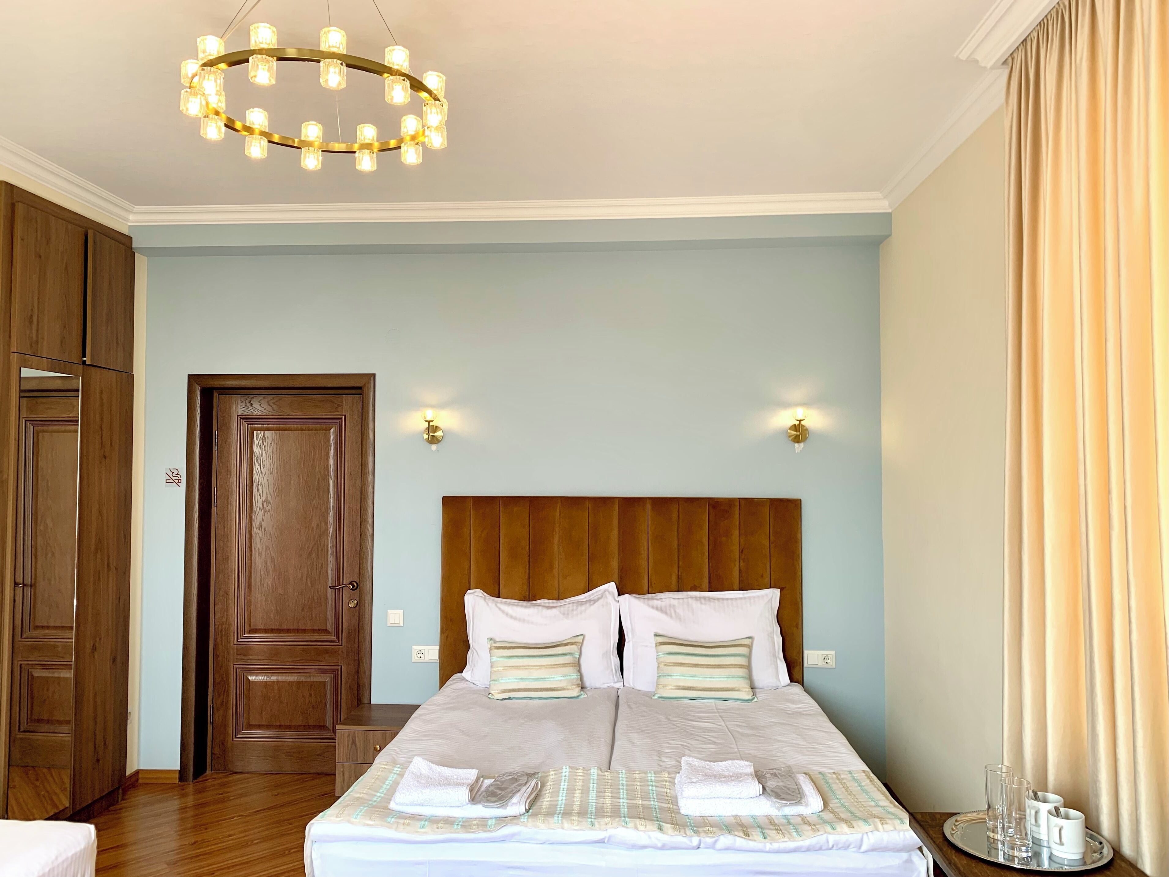 A hotel room with a wooden headboard, a bed with white linens, a chandelier, and a door with a wooden panel.