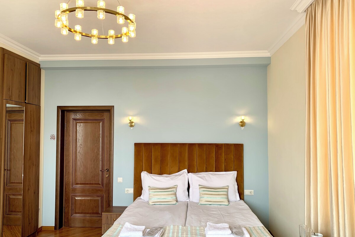 Une chambre d’hôtel avec une tête de lit en bois, un lit recouvert de draps blancs, un lustre et une porte ornée d’un panneau en bois.