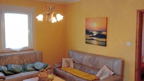 Ein Wohnzimmer mit einer Couch, einem Couchtisch und einem Sessel. Ein Fenster mit Vorhängen, ein Gemälde an der Wand und eine Chandeliebre sind zu sehen.