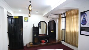 A hotel room with a bed, a vanity with a mirror, a red vacuum cleaner, a black door labeled "SHIVA", and a window with curtains.