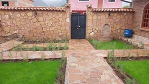 Un patio con muros de piedra y un sendero central que lleva a una puerta negra, rodeado de césped verde y plantas pequeñas.