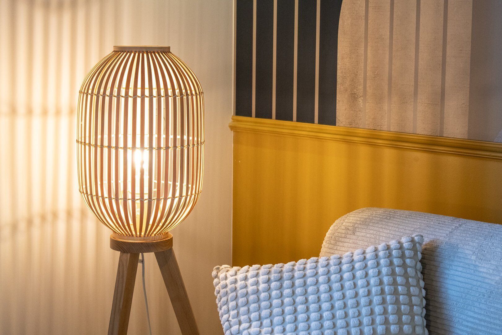 A lamp with a ribbed design on a tripod stand, a textured white pillow, and a yellow wall with vertical stripes.