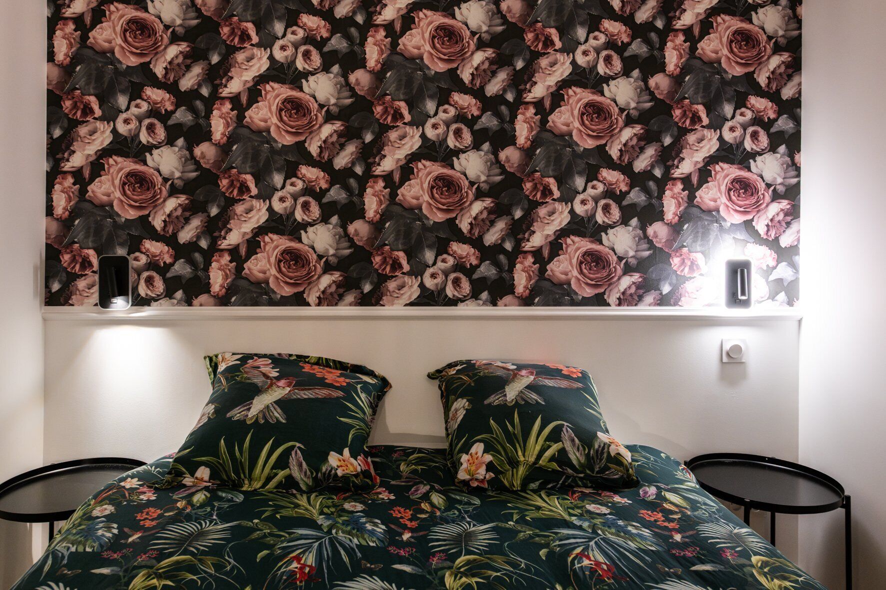 A bedroom with a floral wallpaper, two matching pillows, a bed, and a bedside table.