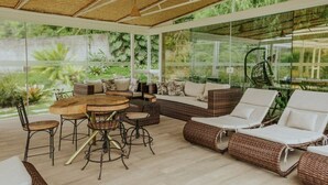 Une vaste terrasse dotée d’une table ronde en bois et de chaises, de canapés en osier et d’une véranda en verre offrant une vue sur une végétation luxuriante.