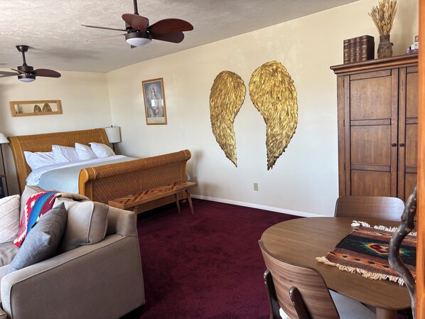 Un dormitorio con cama, sofá, mesa de comedor, ventilador de techo y una pared con alas doradas.
