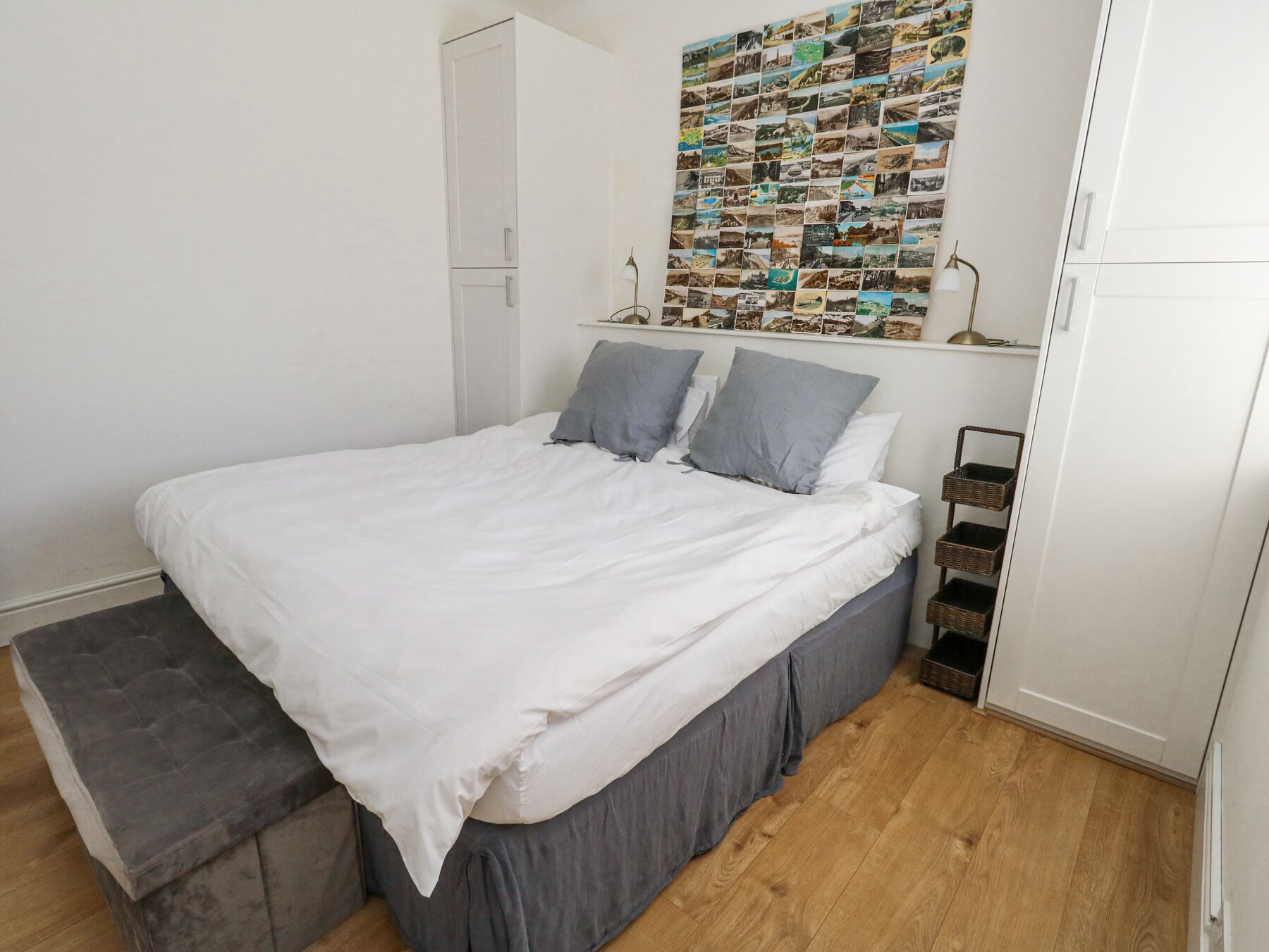 A bedroom with a bed, grey pillows, a white duvet, a wooden floor, a grey bench, a wall with a collage of photos, and a white wardrobe.