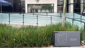 A modern building with a glass facade, a sign indicating 'TO DAY' and '2004', and a palm tree in the foreground. - Today Pinheiros NR (Sao Paulo)
